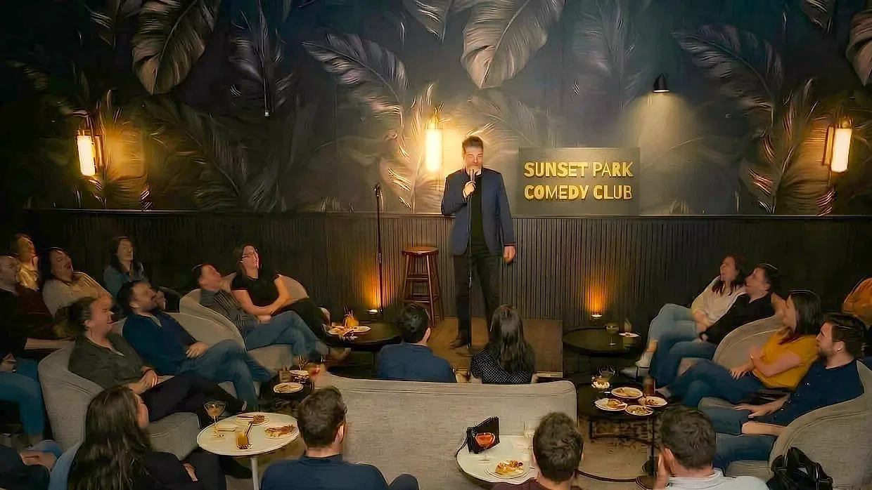 Opening night at Sunset Park Comedy Club: a comic on stage under the club sign, a full room of people laughing at L'Wren